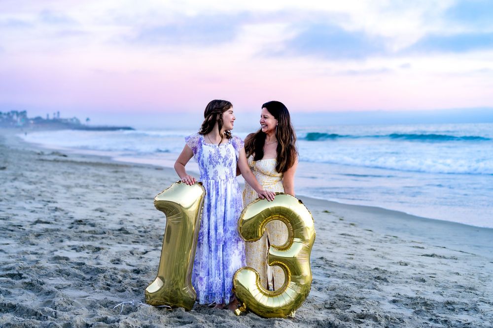 Two people stand on a beach at sunset holding large gold number balloons forming 13.
