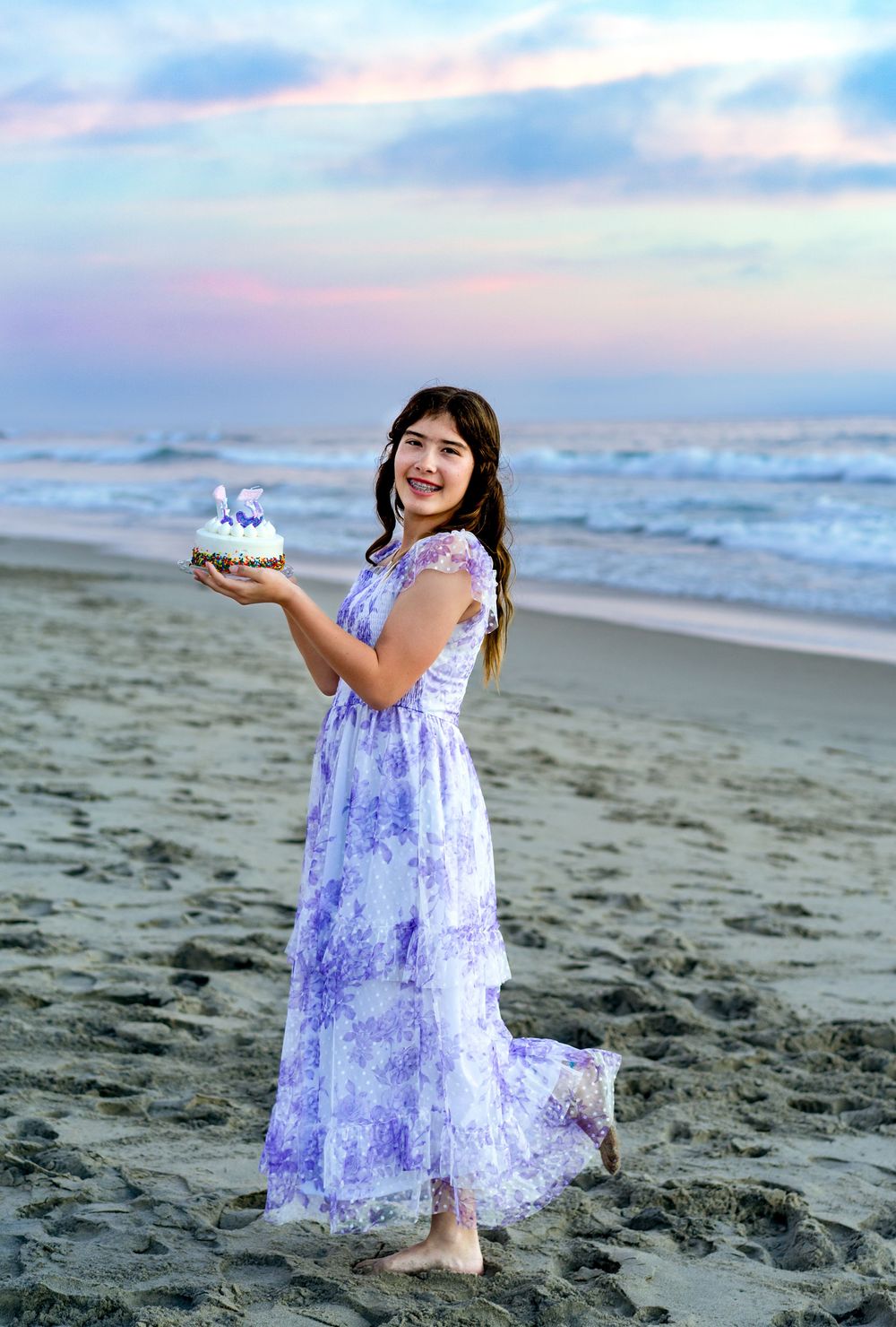 A person in a purple lace dress twirls joyfully on a sandy beach at sunset with waves in the background.