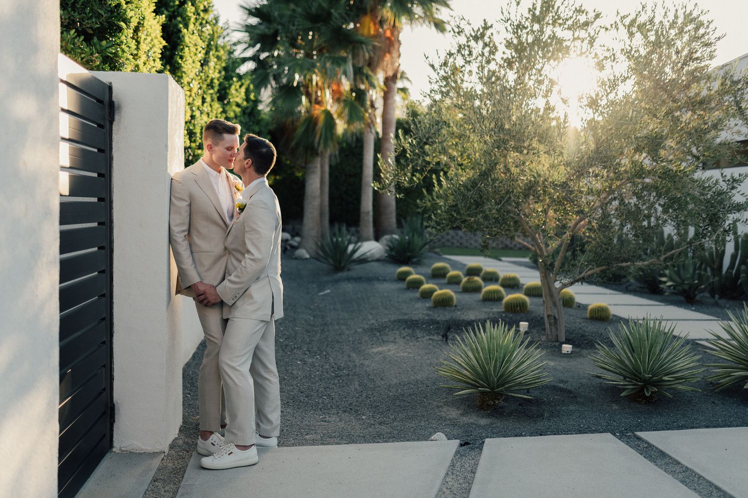 A couple in light gray suits embrace at sunset in a modern desert landscape garden with palm trees and succulents.