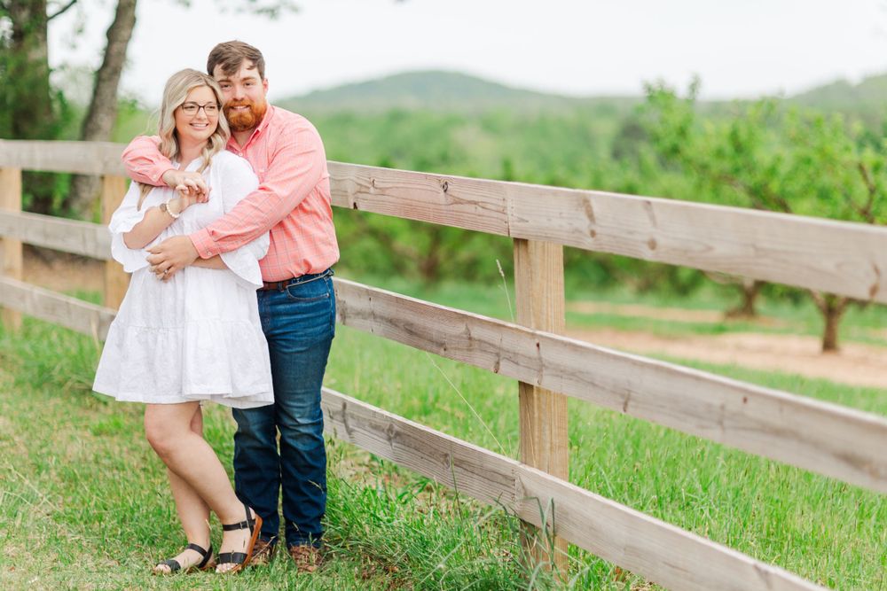 A couple poses against a wooden fence in a rustic countryside setting.