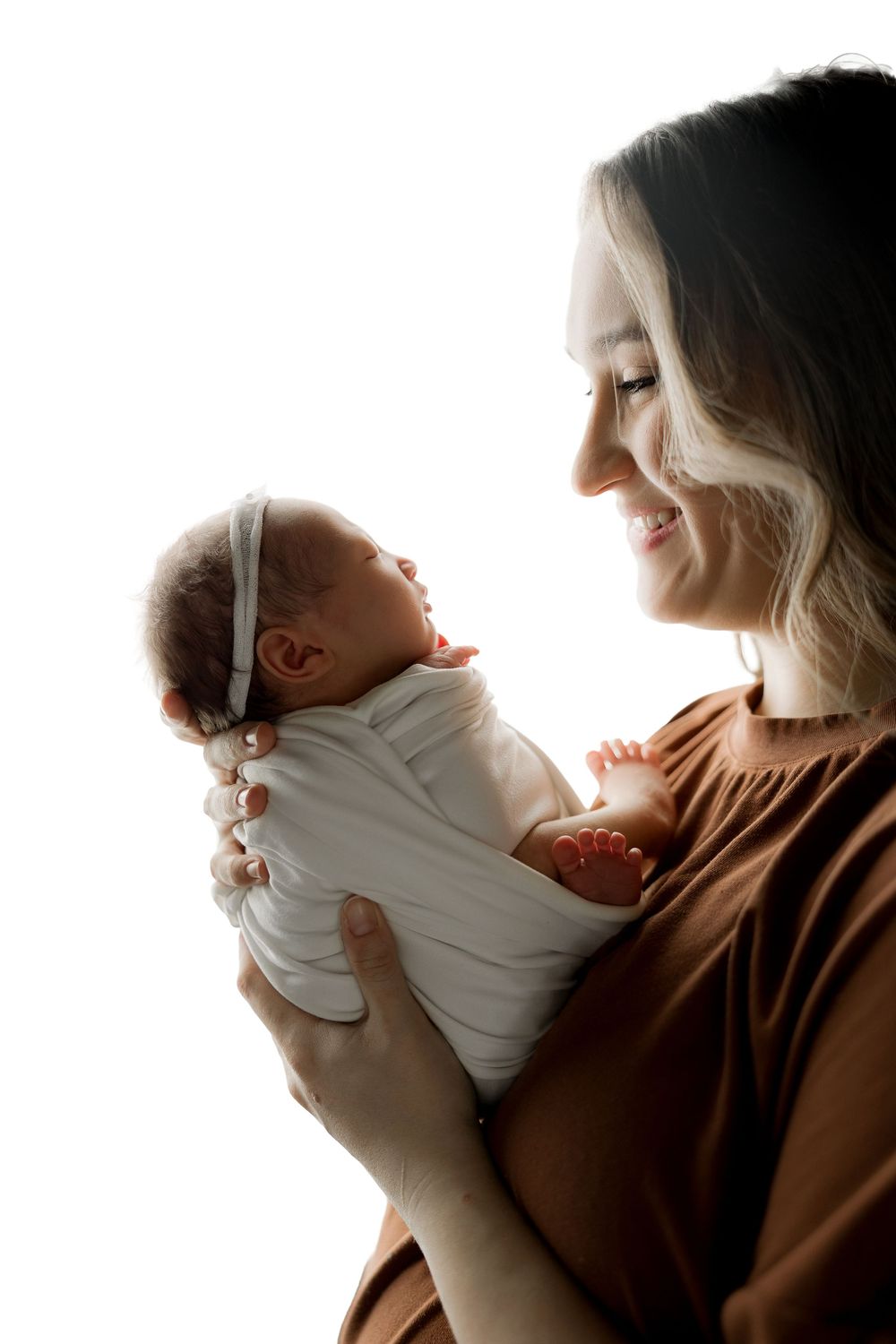 Mother in brown sweater holding newborn baby wrapped in white, illuminated by gentle window light.