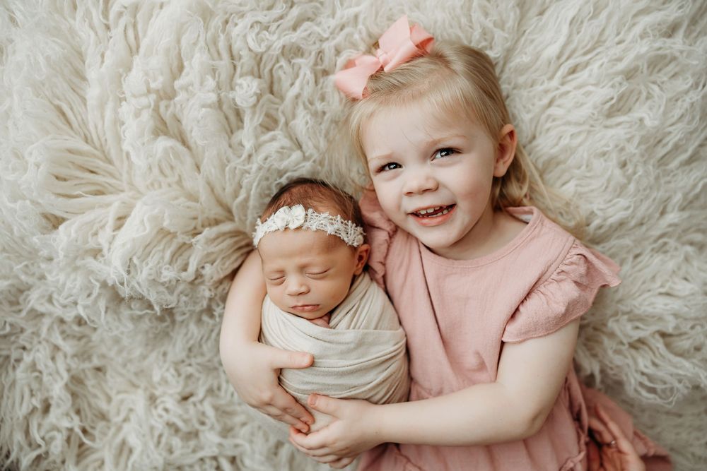 Two young children in matching pink outfits lying on a fluffy white rug while smiling at the camera.