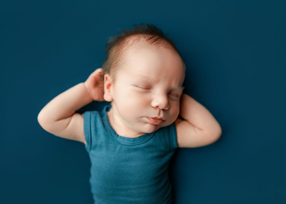 Sleeping newborn baby wearing teal onesie lies peacefully with hands behind head on dark teal background.