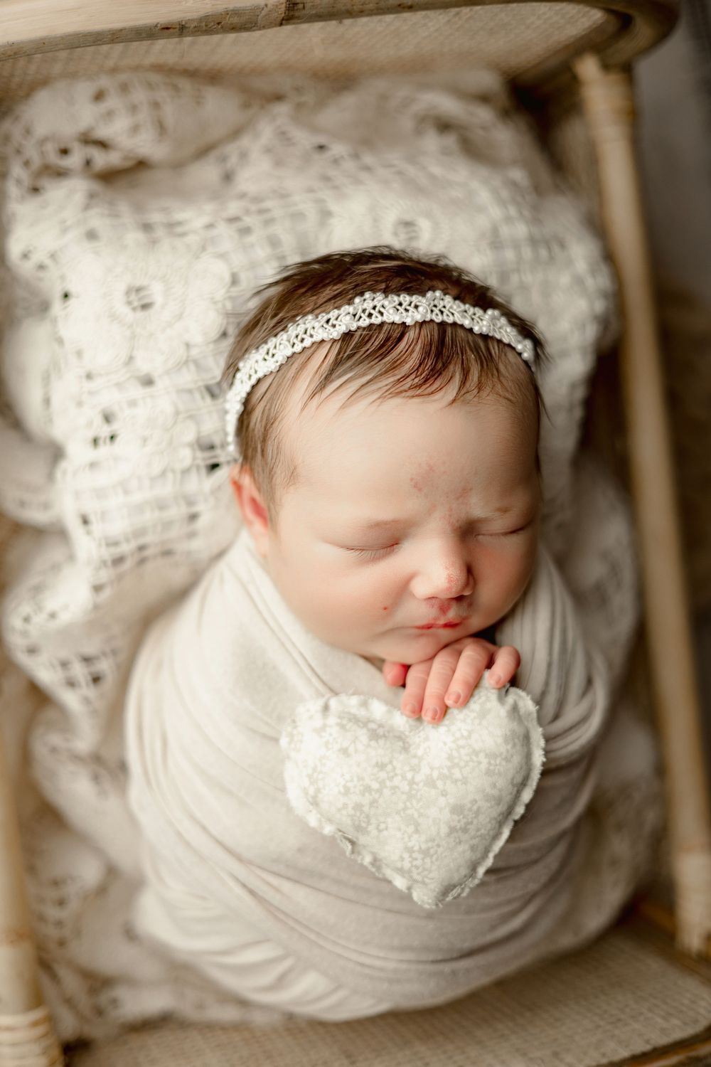 Newborn in white wrap with lace headband resting on textured background with heart decoration.