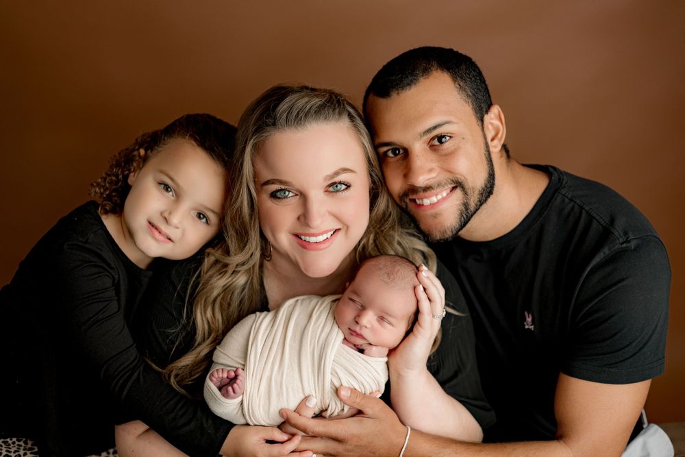 Family portrait against brown background with newborn in white wrap.
