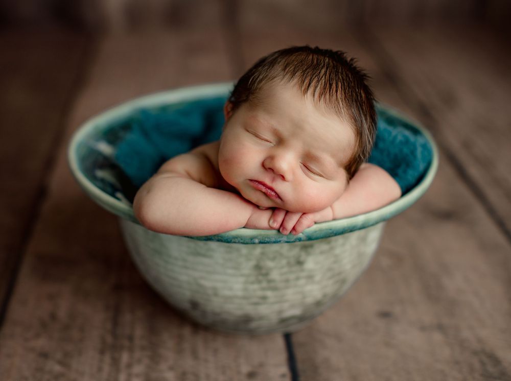 Sleeping newborn peacefully resting in turquoise ceramic bowl on wooden surface.