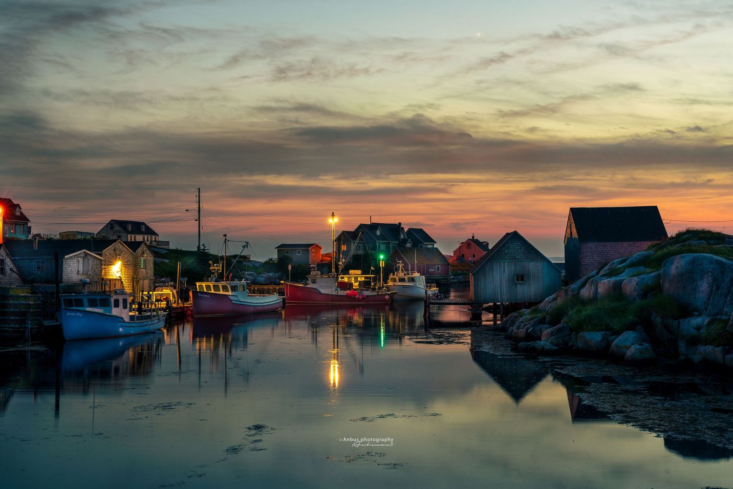 Colorful fishing boats reflected in calm harbor waters at sunset, with rustic waterfront buildings glowing warmly in twilight.