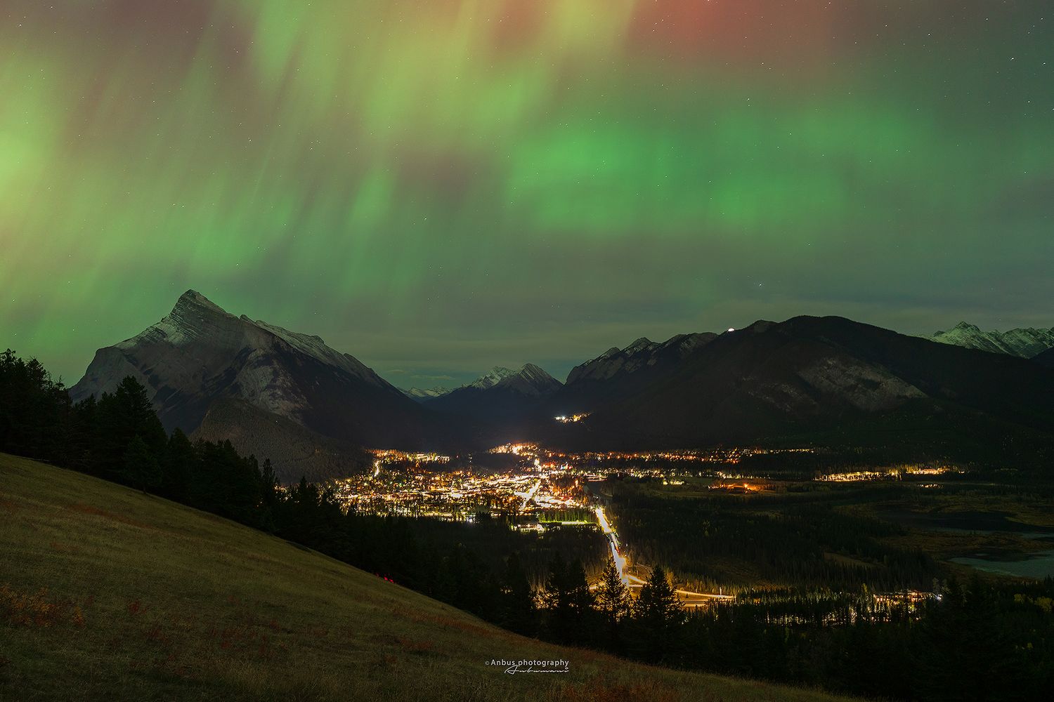 Northern lights glow green above a Banff town with twinkling lights nestled in a valley at night.