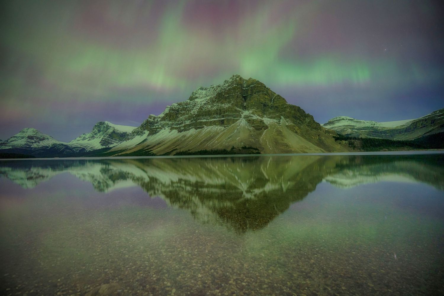 Majestic mountain peak reflecting in calm lake waters under vibrant aurora borealis lights in the night sky.