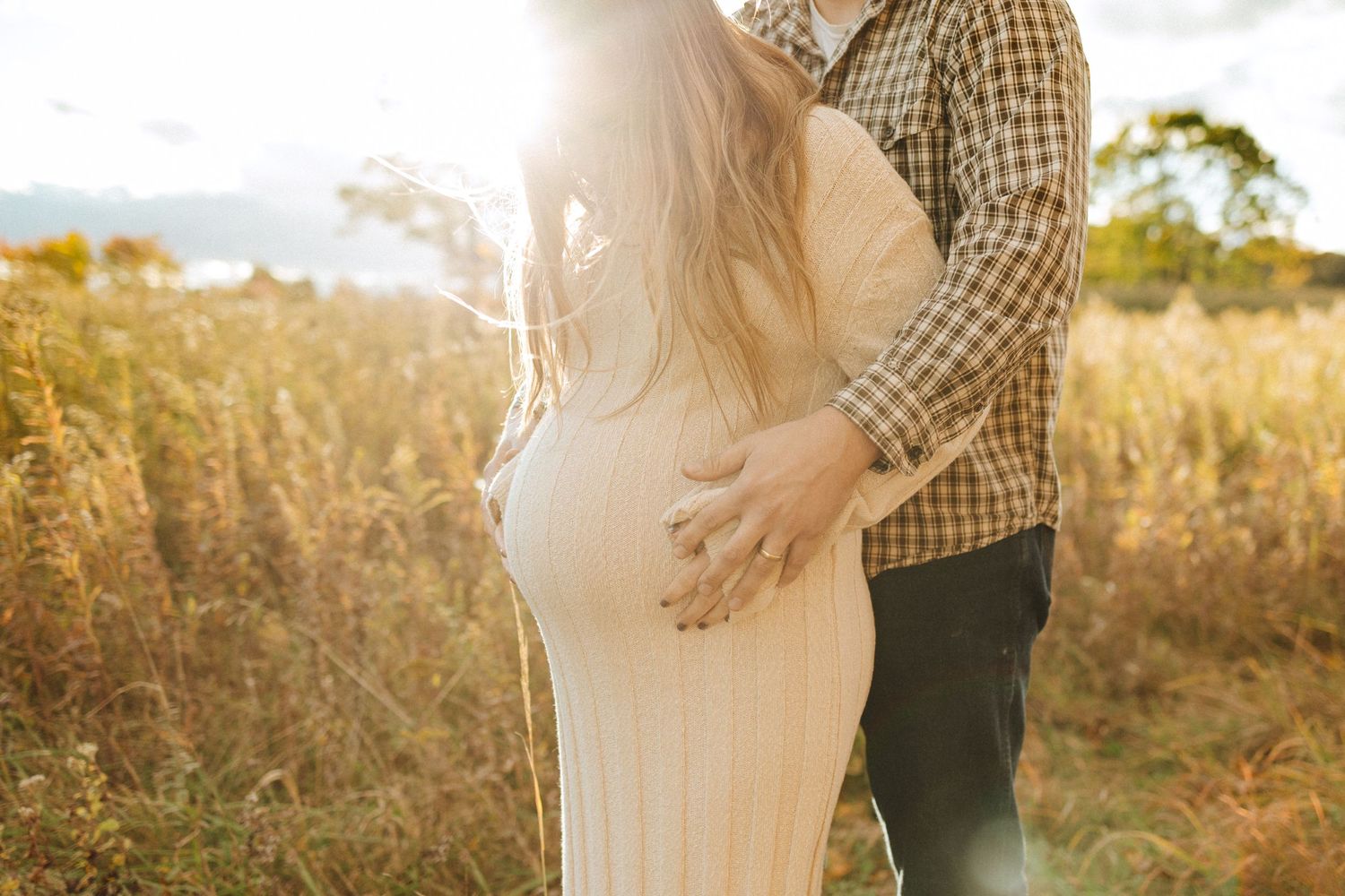 A couple embraces in a sunlit field during golden hour, with the expecting mother wearing a flowing beige dress in califon nj