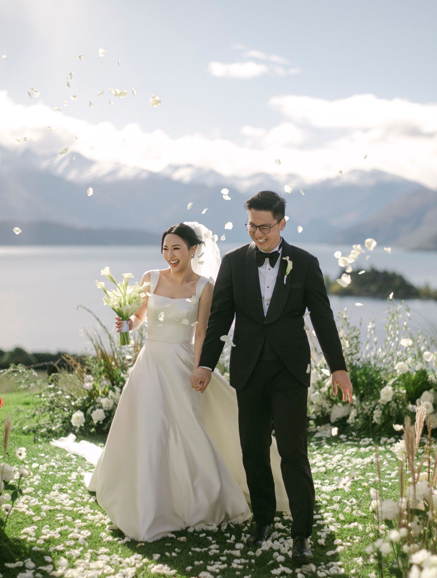 A joyful couple walks hand-in-hand after their outdoor wedding ceremony, with petals falling and a stunning lake and mountain backdrop.