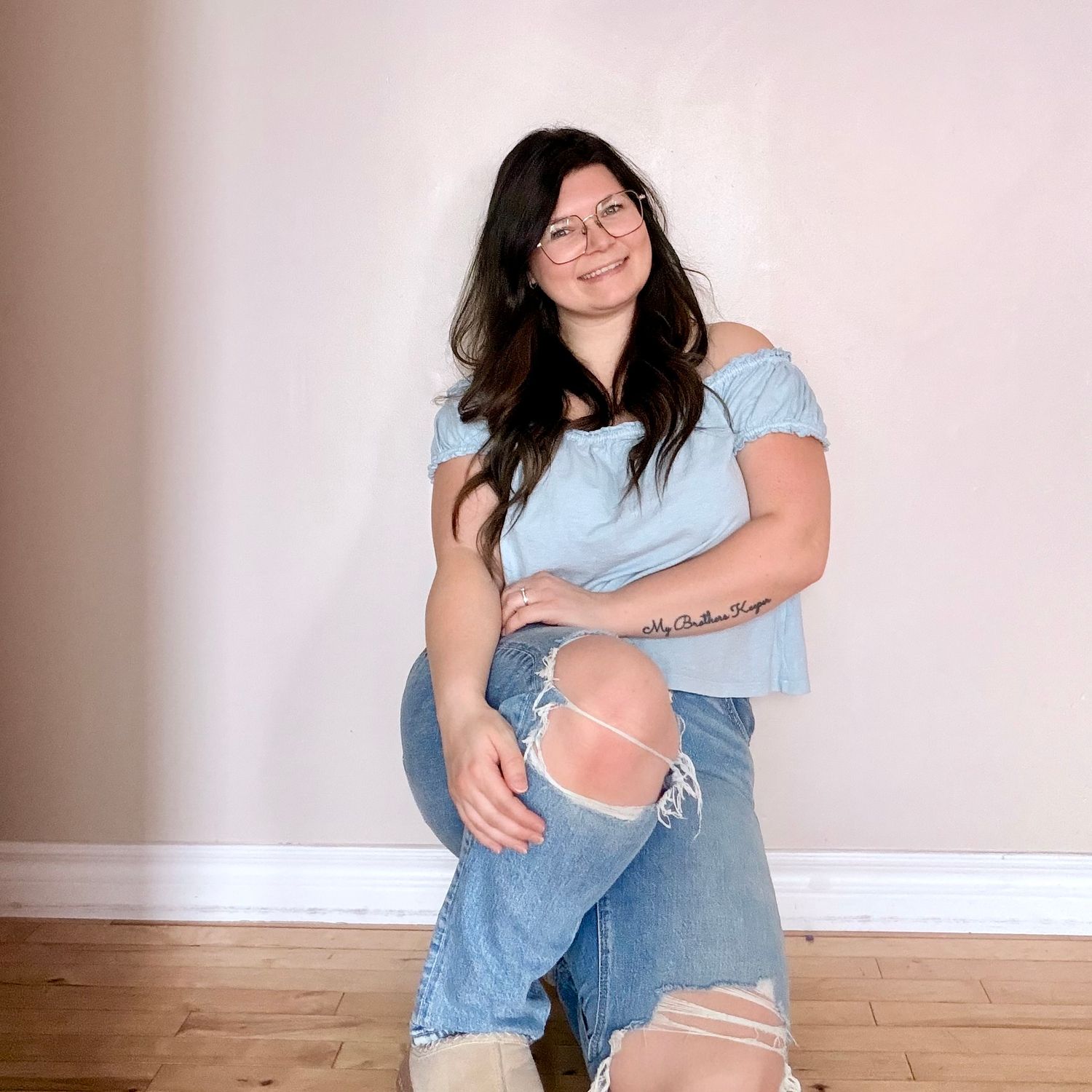 A young woman in ripped jeans and off-shoulder blue top sits casually against a neutral wall indoors.