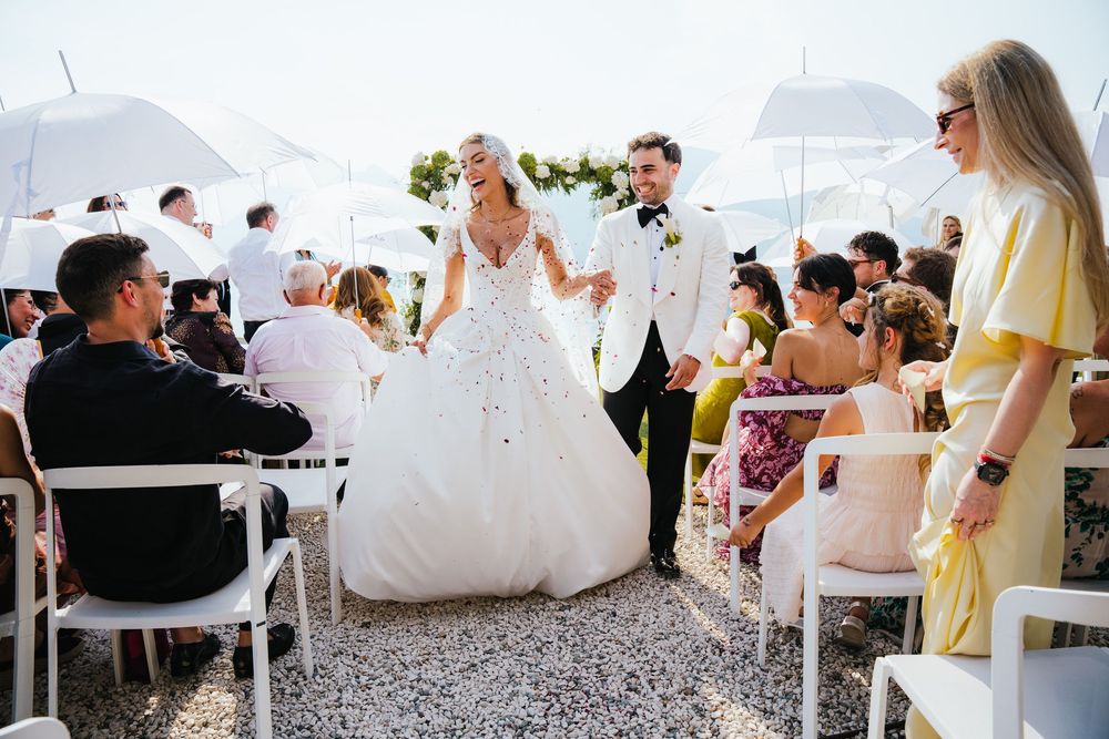 Bride and groom walk down the aisle at an outdoor wedding ceremony with guests seated in white chairs.