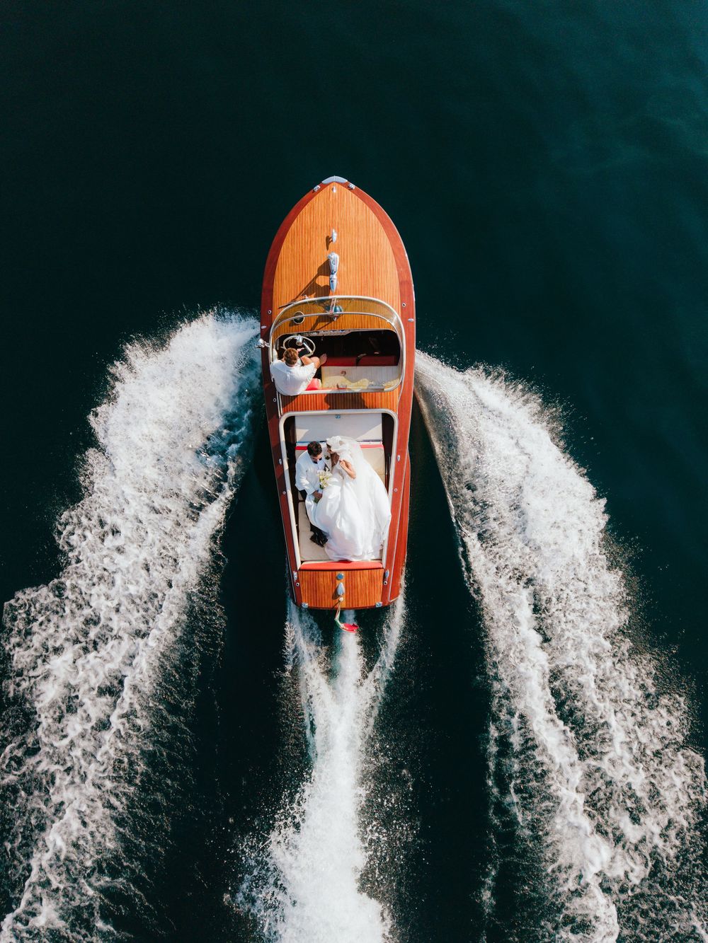 Aerial view of a bride and groom riding a classic wooden speedboat on deep blue water, creating white wake trails.