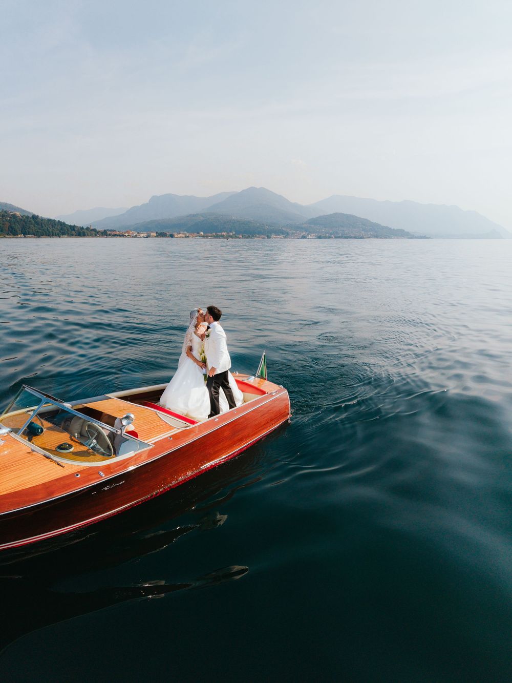 Bride and groom kissing on a classic wooden speedboat on a calm lake with misty mountains in the background.