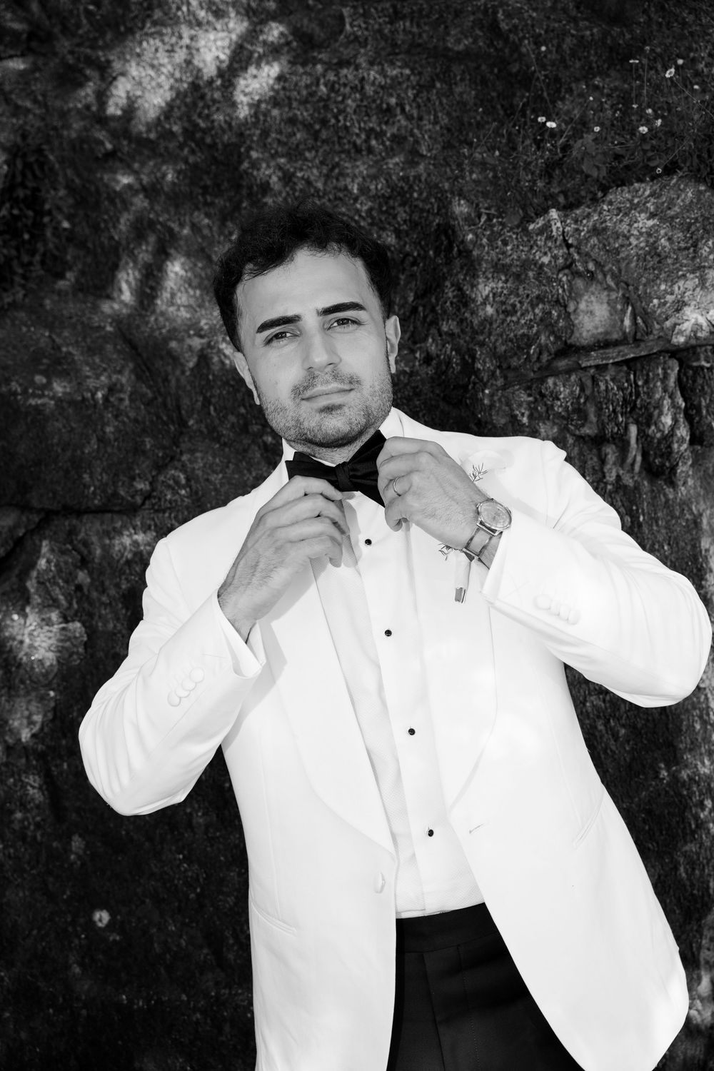 A dapper groom in a white tuxedo adjusts his black bow tie against a stone wall in a classic black and white portrait.