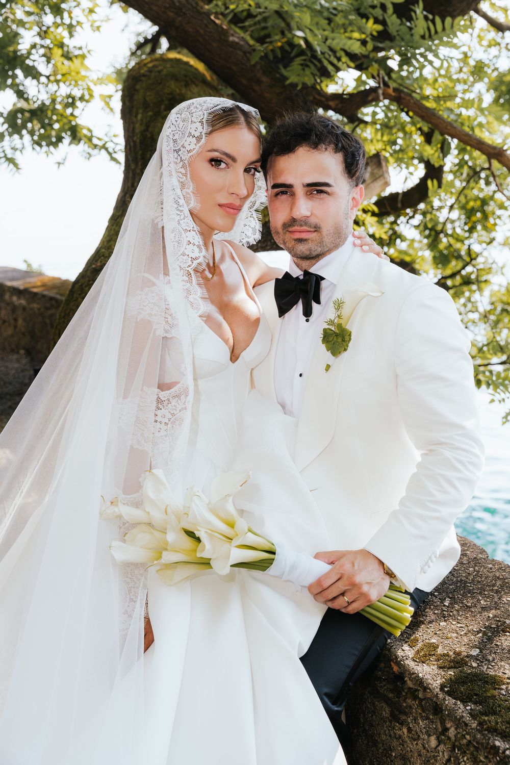 Elegant bride in lace veil holding calla lily bouquet poses with groom in white tuxedo under a tree by the water.
