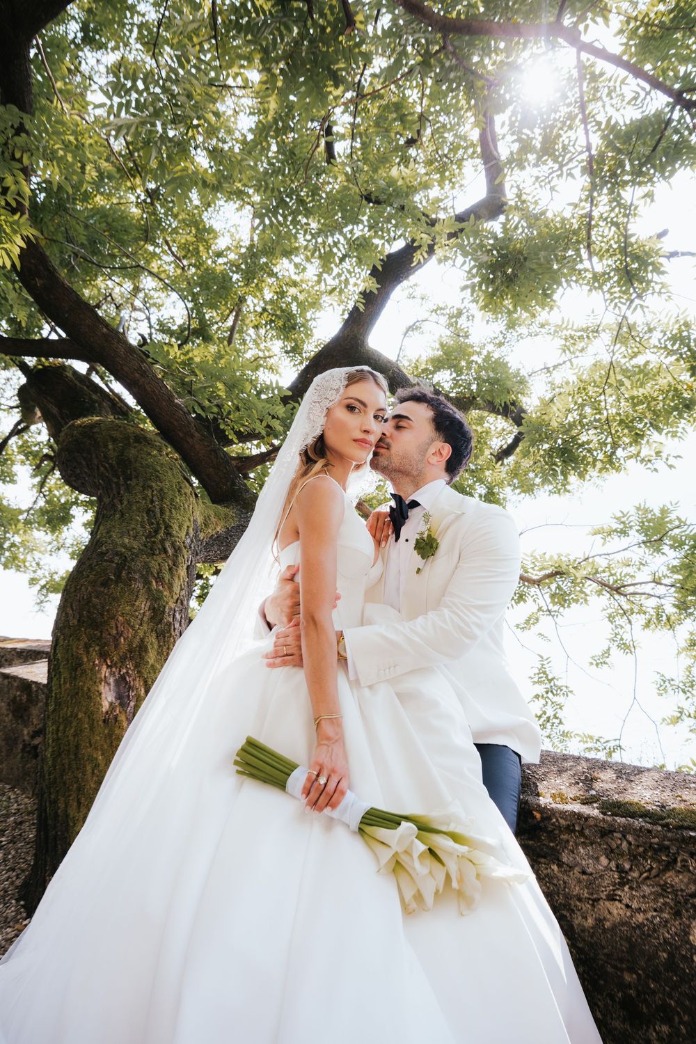 Bride and groom embrace under a large mossy tree, bride holding white calla lily bouquet in elegant wedding attire.