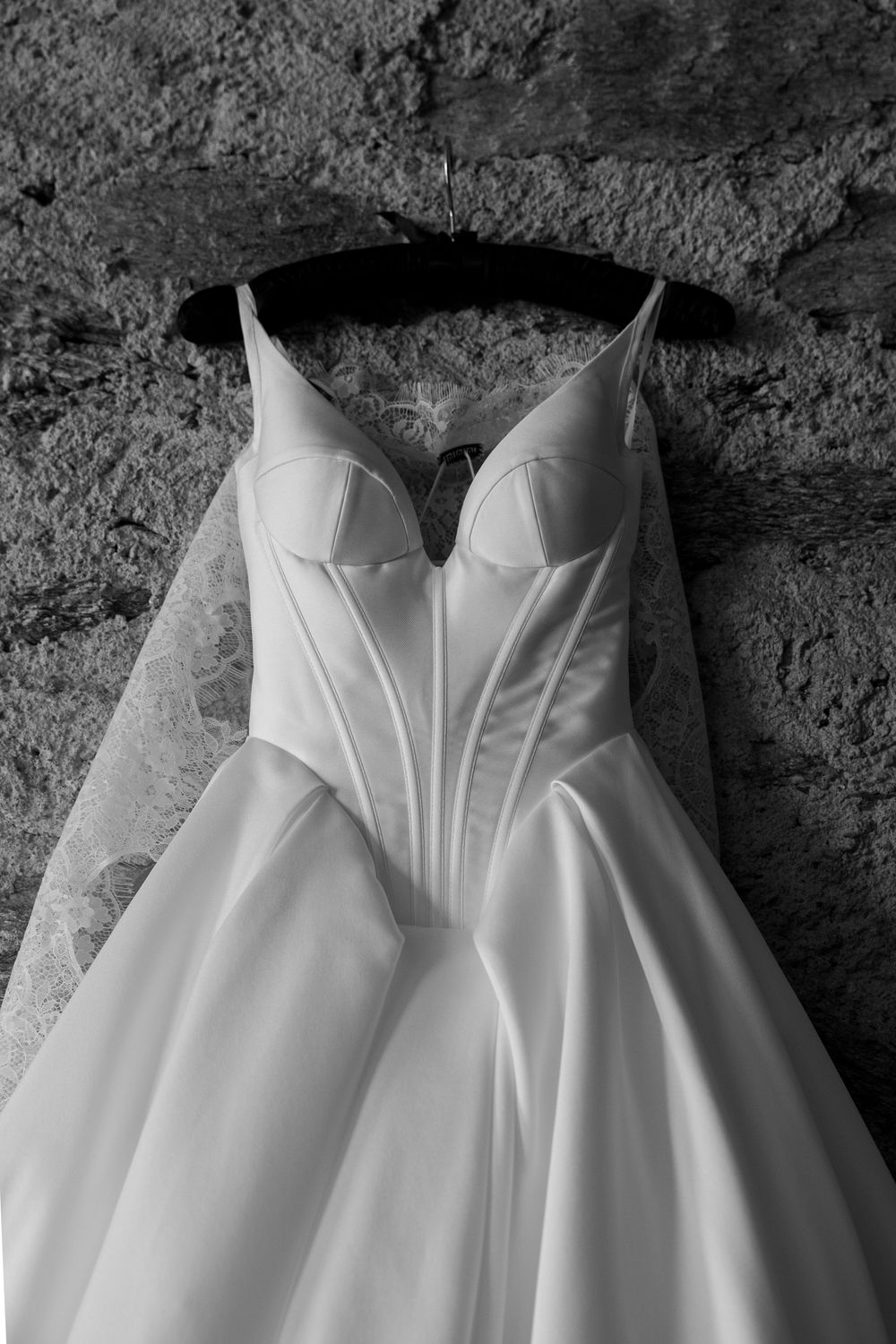 Black and white photo of an elegant satin wedding gown with corset bodice hanging on a hanger against a stone wall.