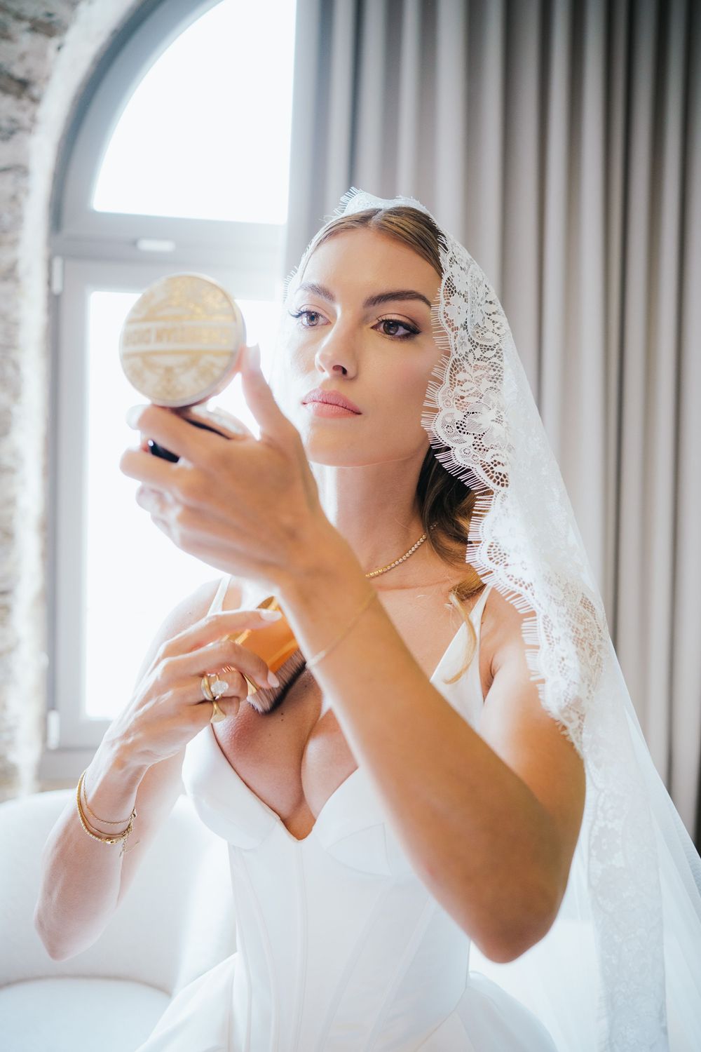 Bride in white gown and lace veil checks makeup in compact mirror, wearing gold jewelry in bright room.