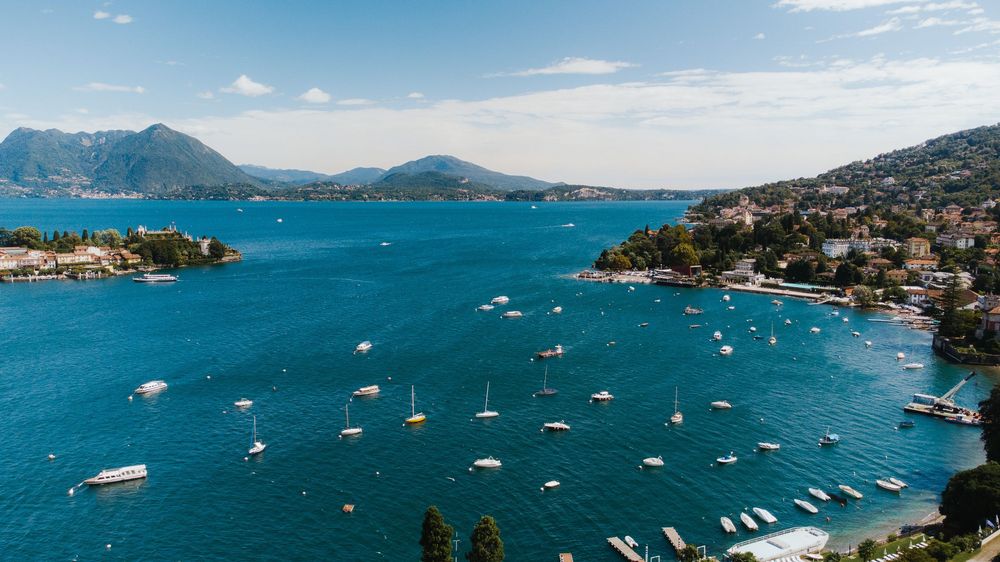 Aerial view of a scenic Italian lake with sailboats, lush hillside towns, and mountains under a bright blue sky.