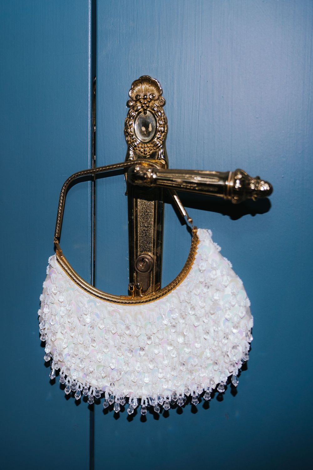 White beaded bridal clutch purse hanging on ornate gold door handle against teal blue wall.