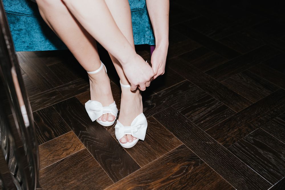 Detail shots of bride putting on white lace wedding shoes while sitting down.