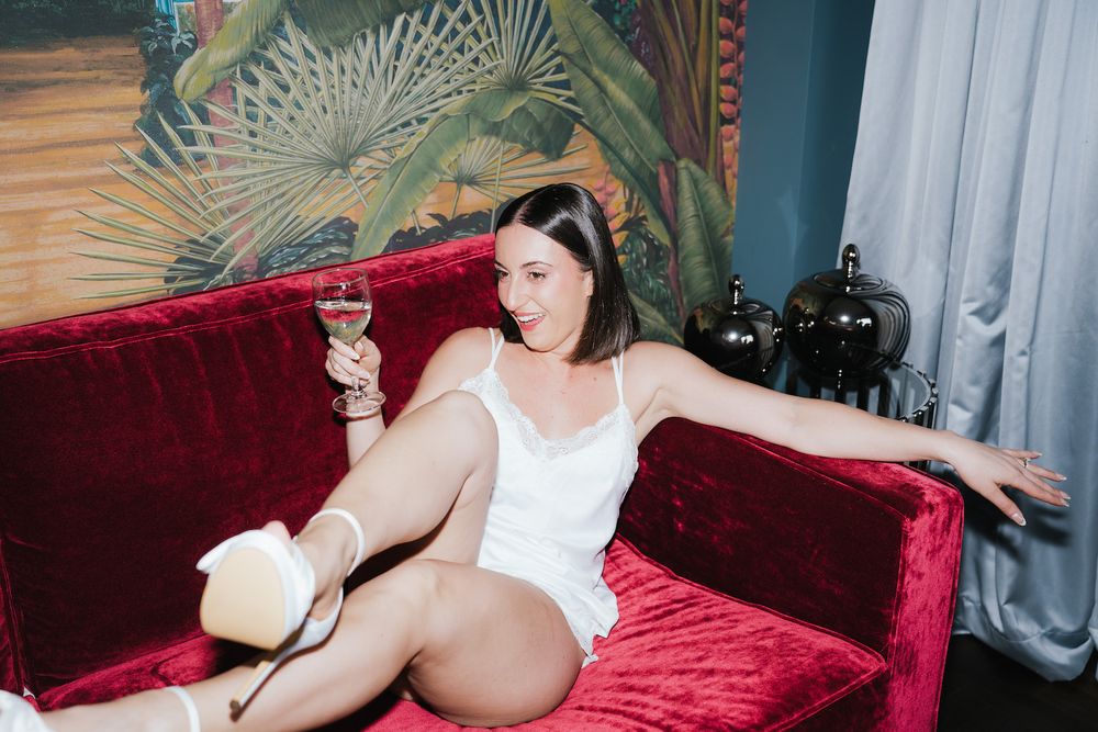 Woman in white lingerie lounges on red velvet couch holding champagne glass.