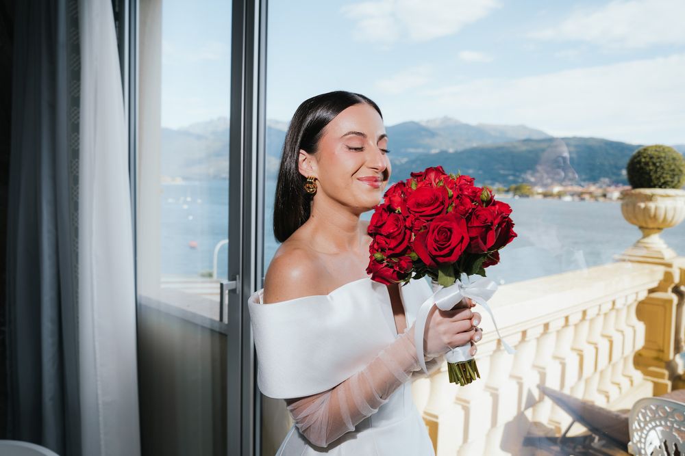 Bride holding red rose bouquet on balcony overlooking coastal mountain landscape.