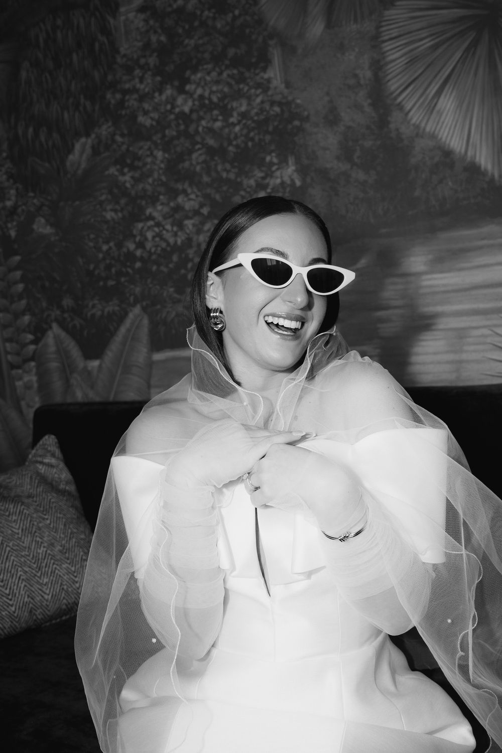 Black and white photo of bride in retro sunglasses and structured white dress laughing.