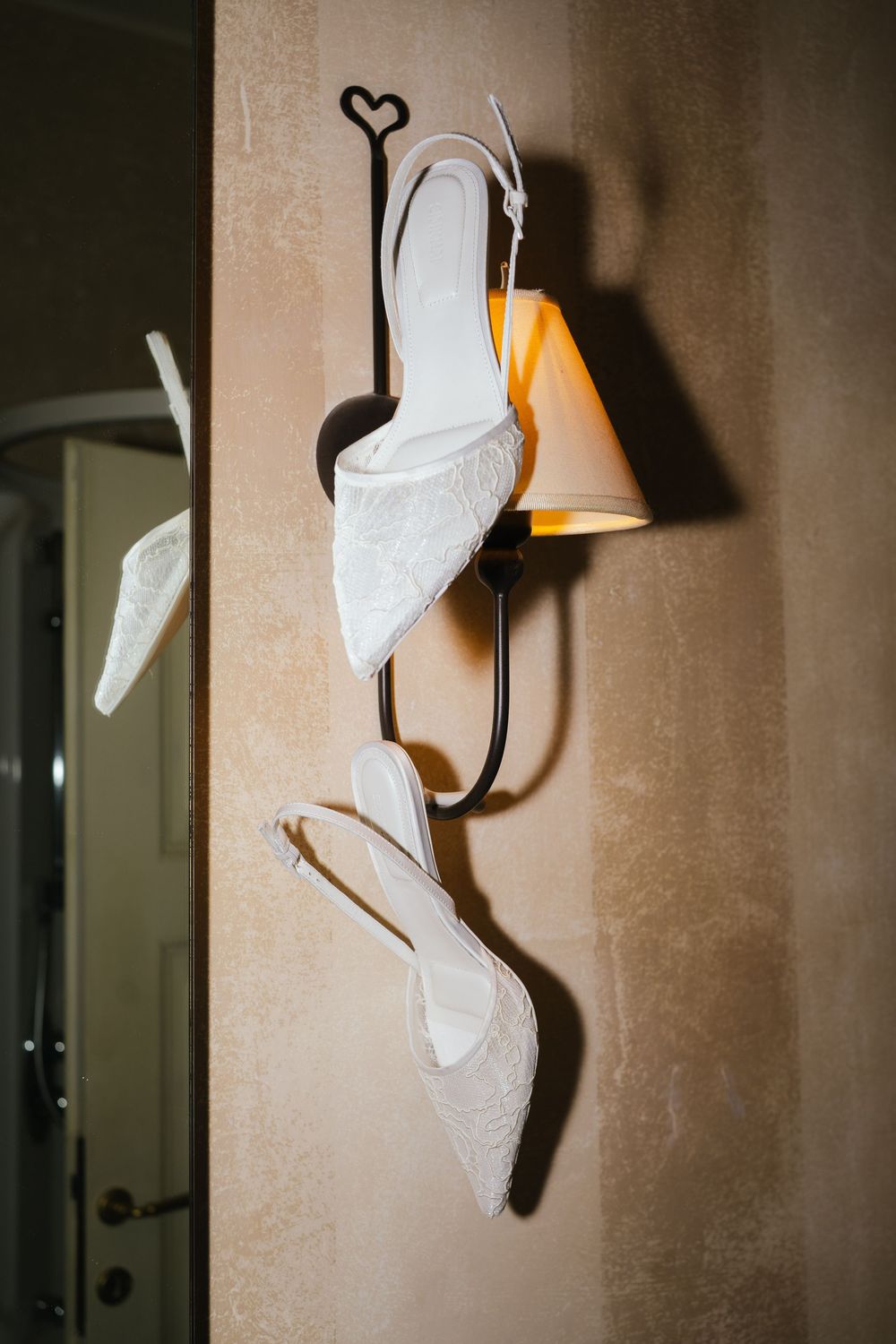 Elegant white lace bridal heels hanging on a decorative wall sconce with warm golden light.