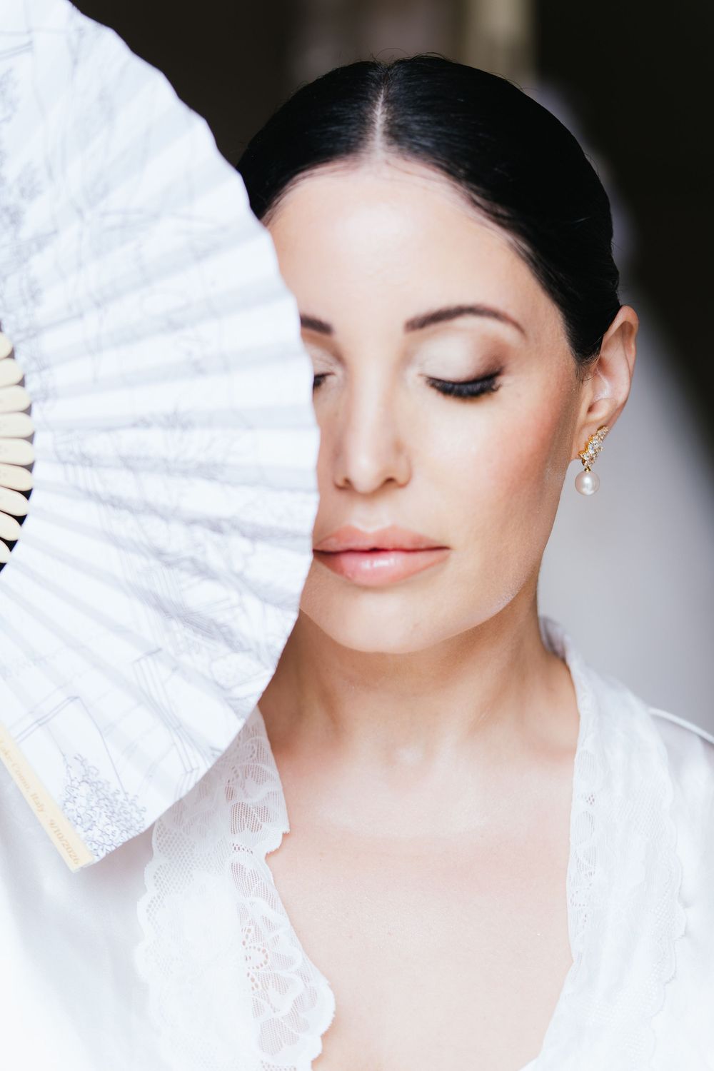 Elegant bride in white lace robe holding a decorative white fan near her face with eyes closed.