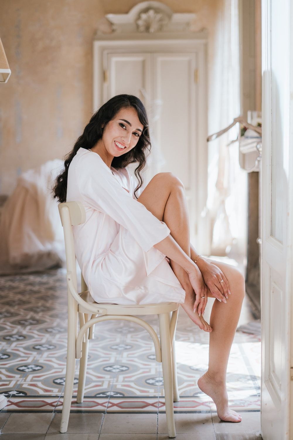 Woman in white robe sits on vintage chair in sunlit room with patterned tile floor.