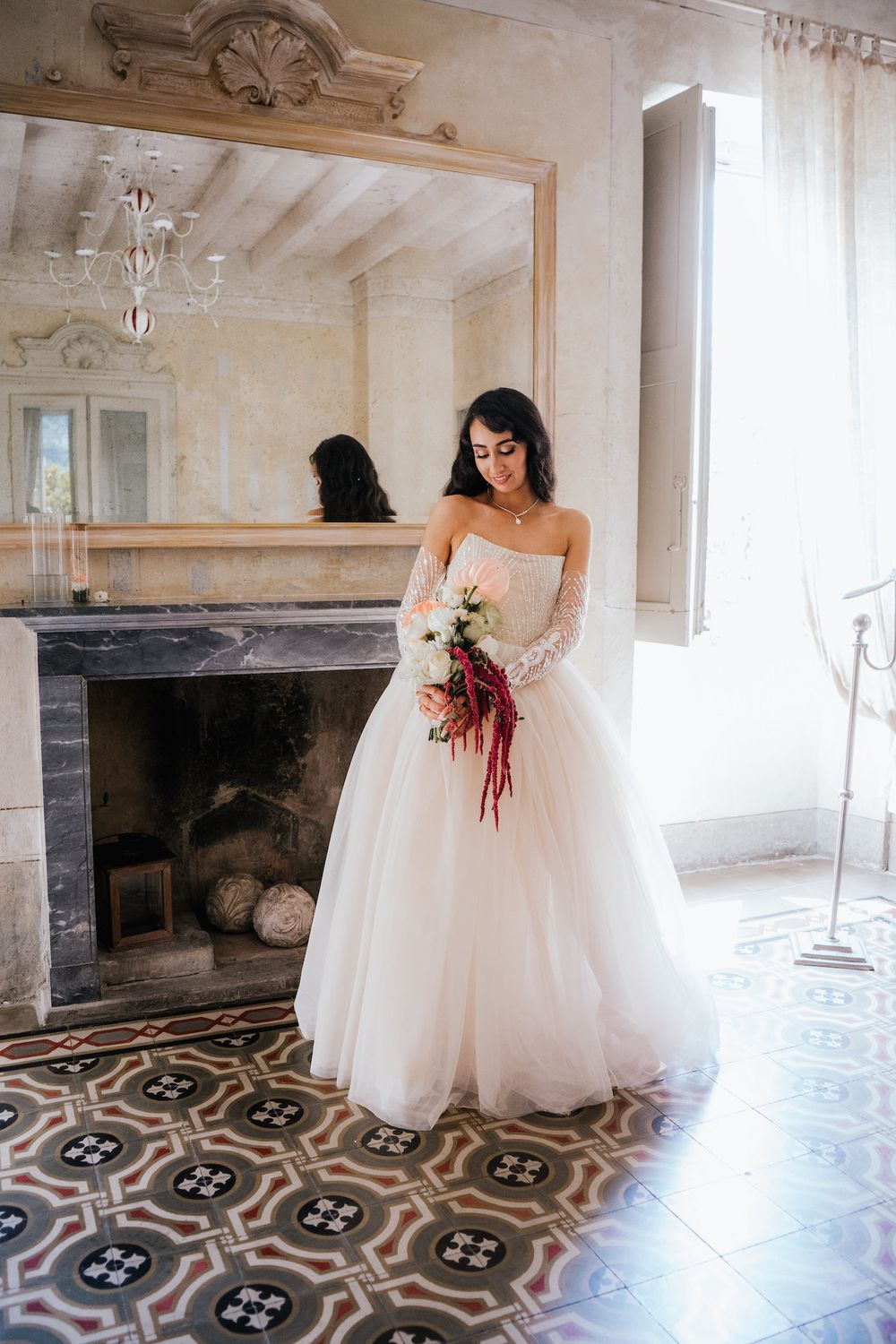 Bride in strapless ballgown holds burgundy bouquet by ornate fireplace.
