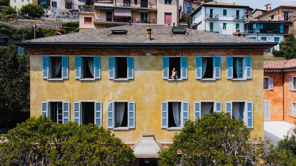 Yellow Italian villa with blue shuttered windows and a tiled roof in a hillside setting.