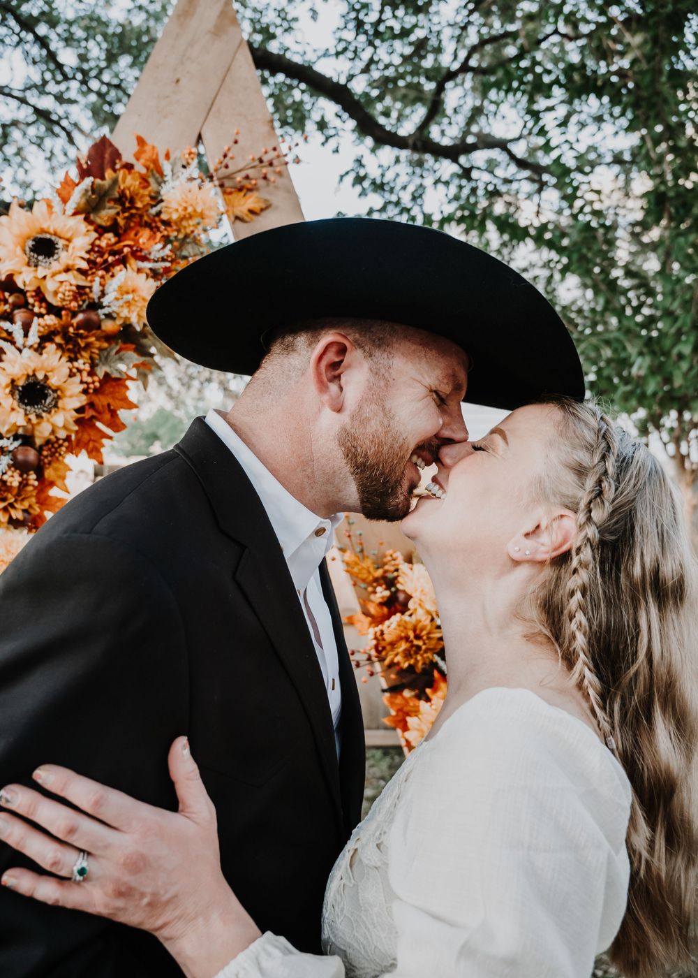 Couple sharing a kiss at fall wedding ceremony under floral arch with sunflowers and autumn blooms.
