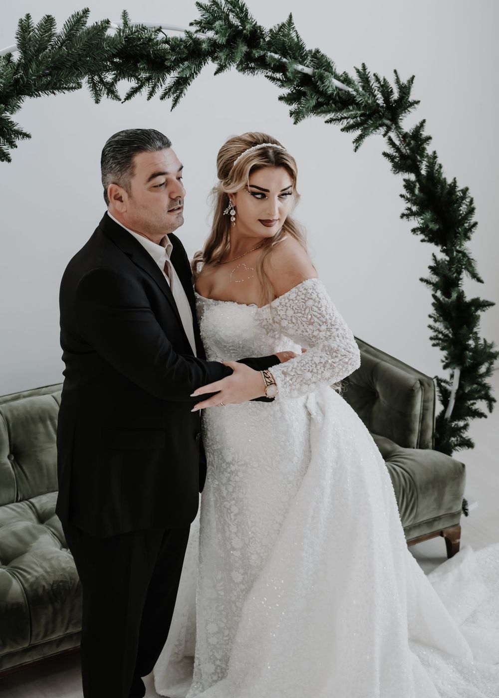Couple poses in wedding attire under pine garland arch, bride in off-shoulder lace gown, groom in black suit.