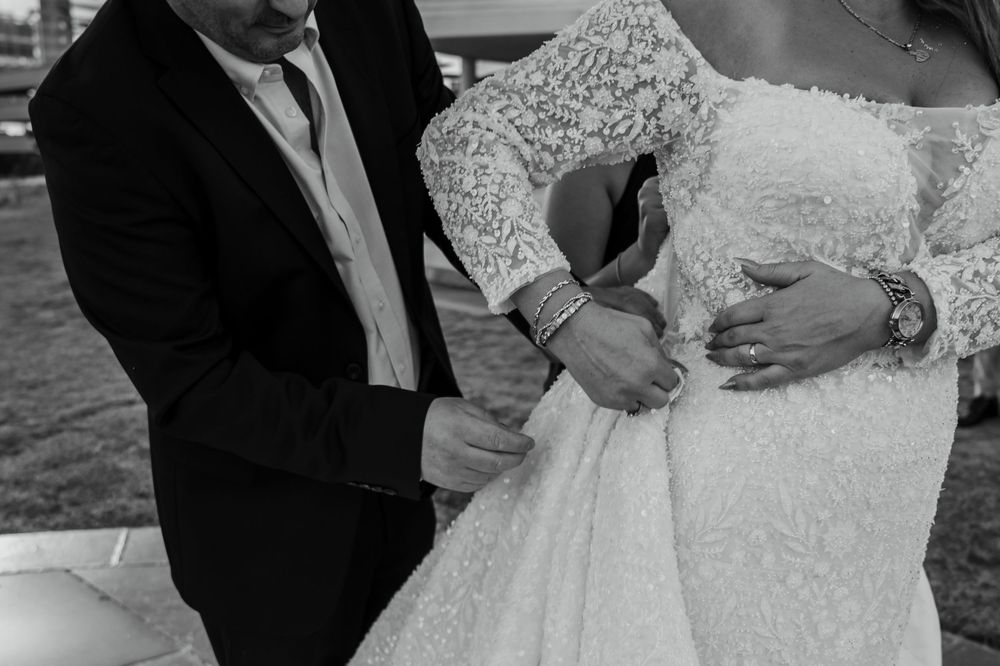 Black and white photo of a bride and groom, groom fastening the back of brides lace wedding dress.