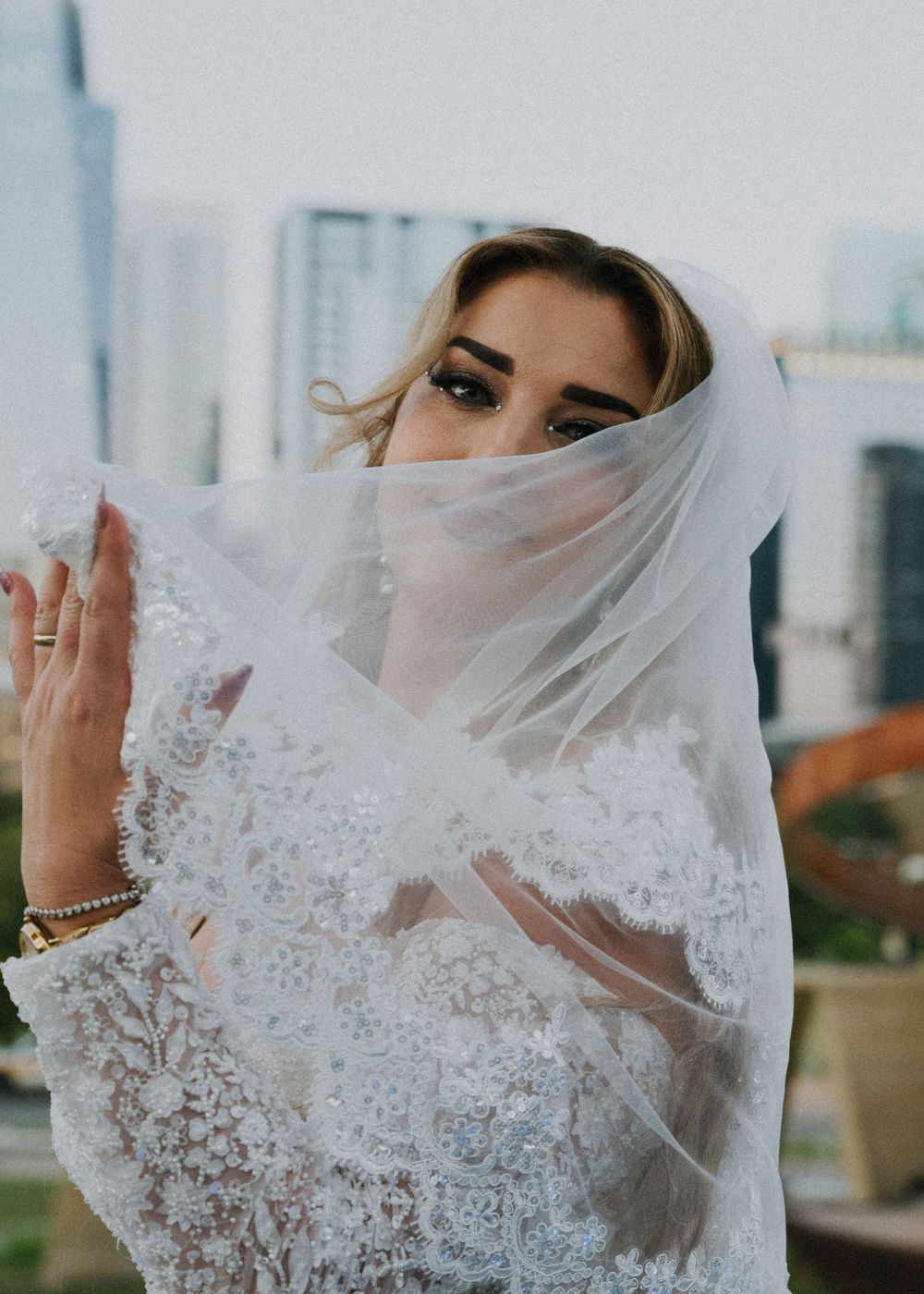 Bride in lace wedding dress with veil draped over face, posing against city skyline backdrop.