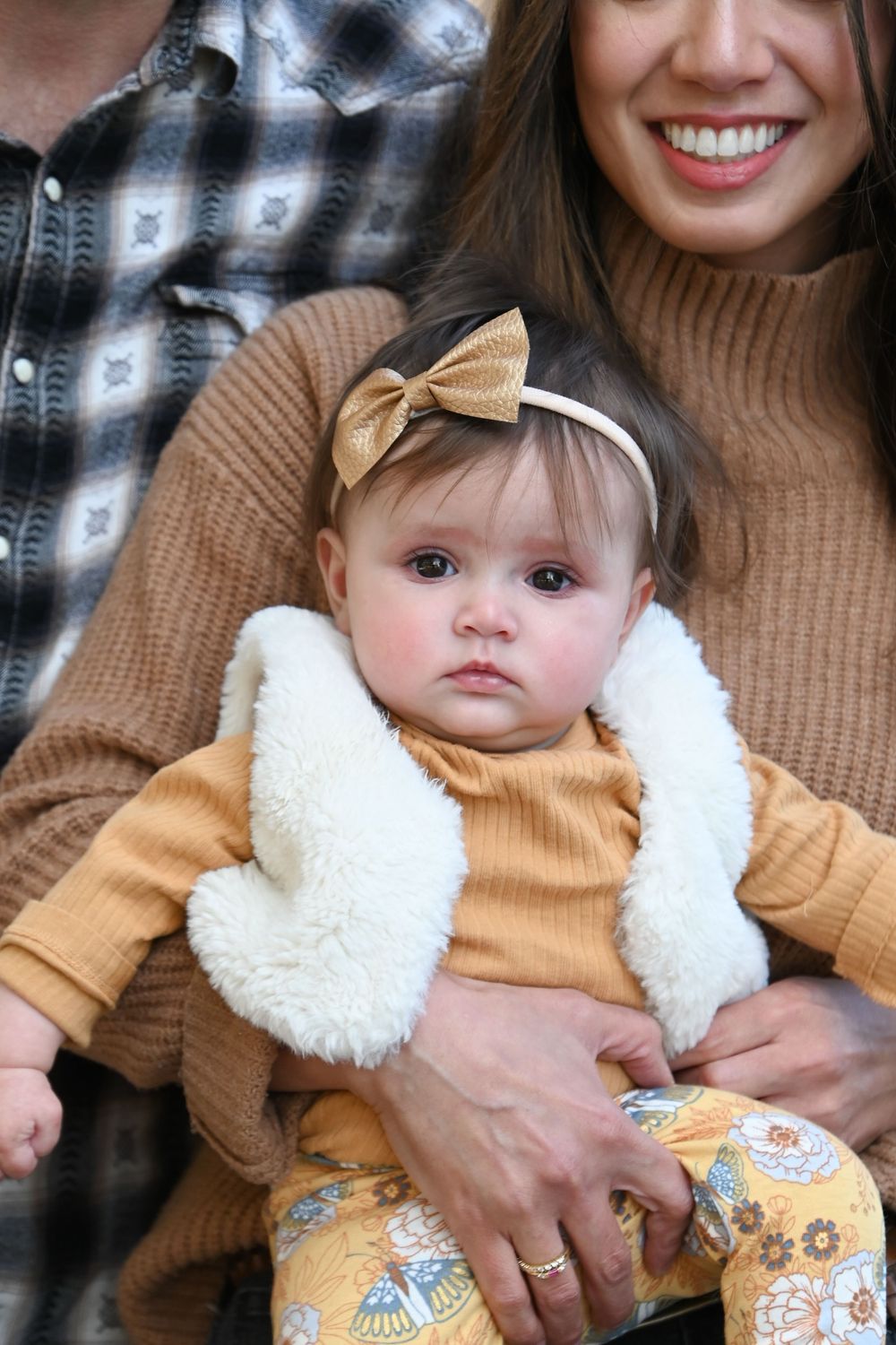A baby in a tan sweater and fuzzy white vest with a gold bow headband sits on a lap with a plaid blanket nearby.