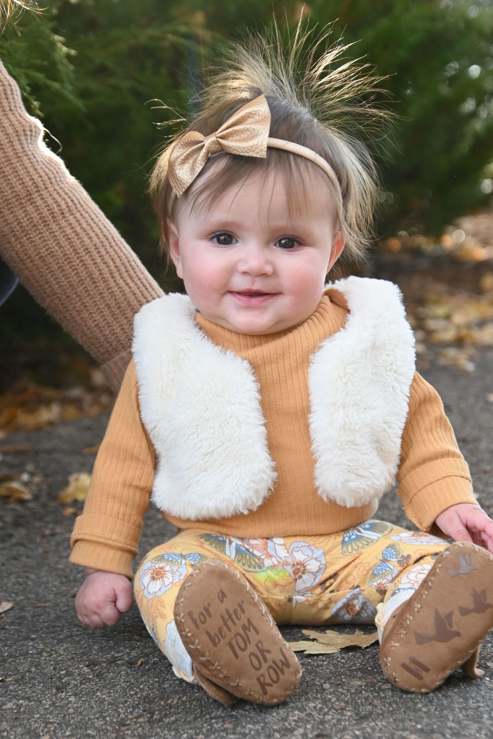 A baby wearing a cozy yellow sweater and white vest sits on autumn leaves with brown shoes.