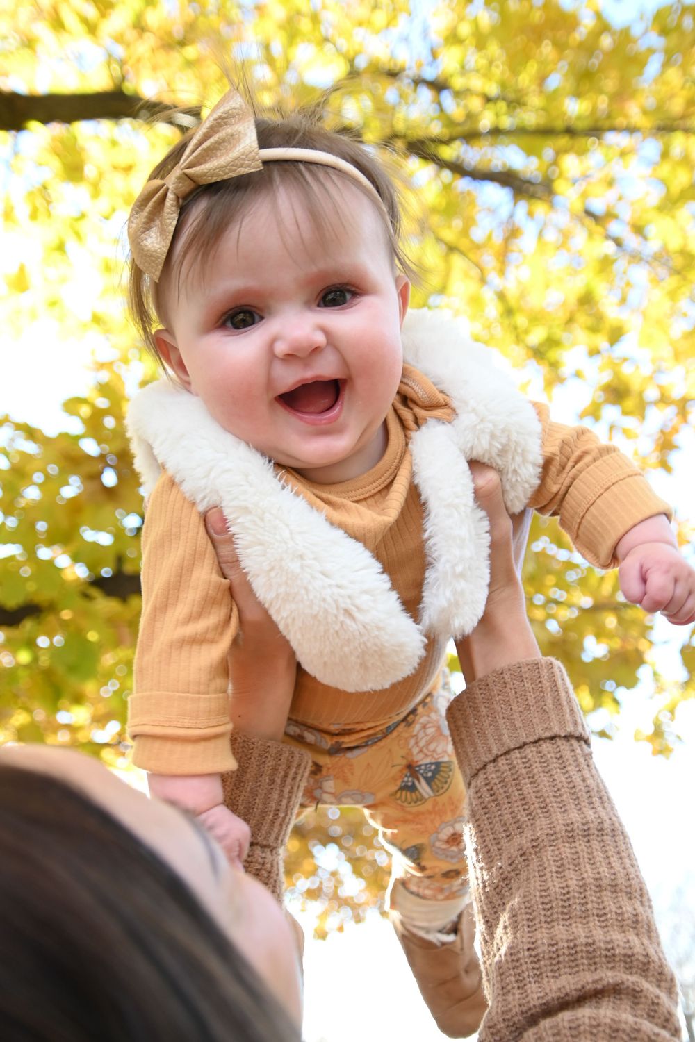 A small baby wearing a cozy tan outfit with white fur collar and bow smiles joyfully against autumn yellow foliage.