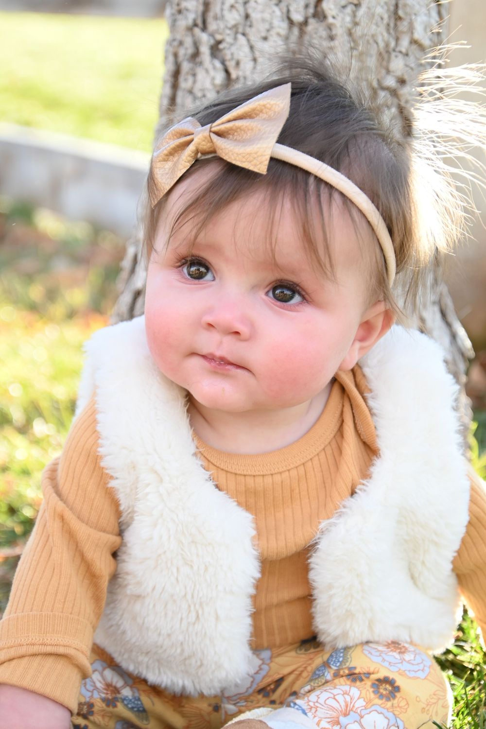 Baby wearing a cozy beige sweater and white furry vest outdoors in autumn scenery.