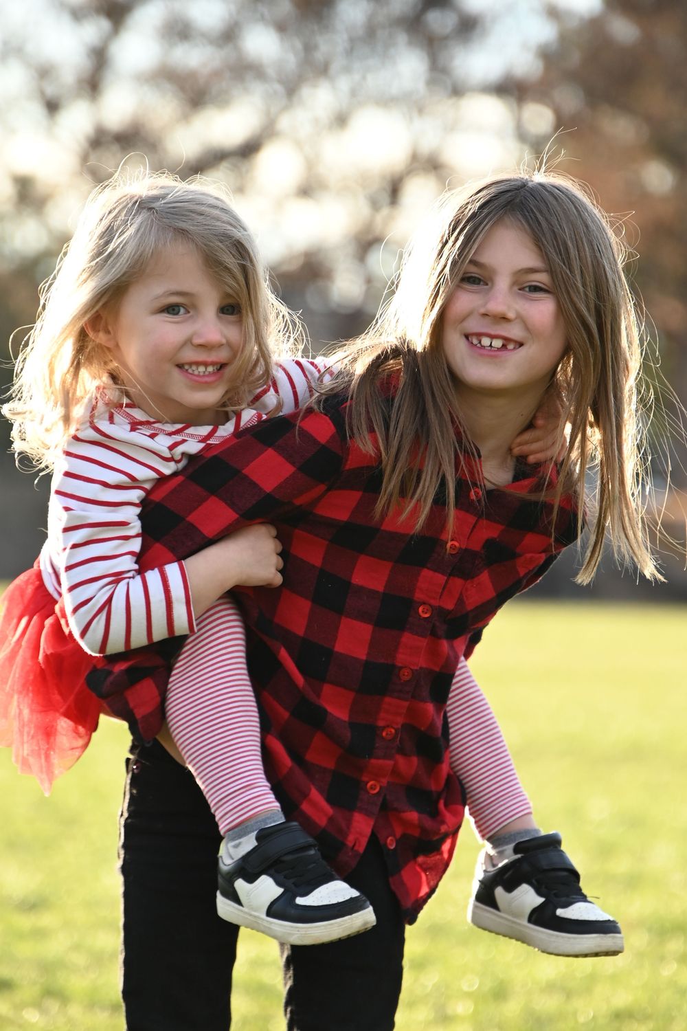 Two children play outdoors at sunset, one wearing a buffalo plaid shirt giving a piggyback ride to the other in stripes.