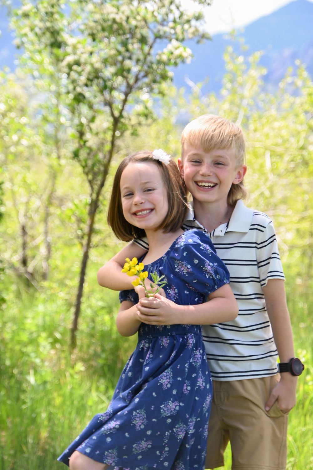 Two children standing together outdoors on a sunny day with yellow wildflowers and green foliage in the background.