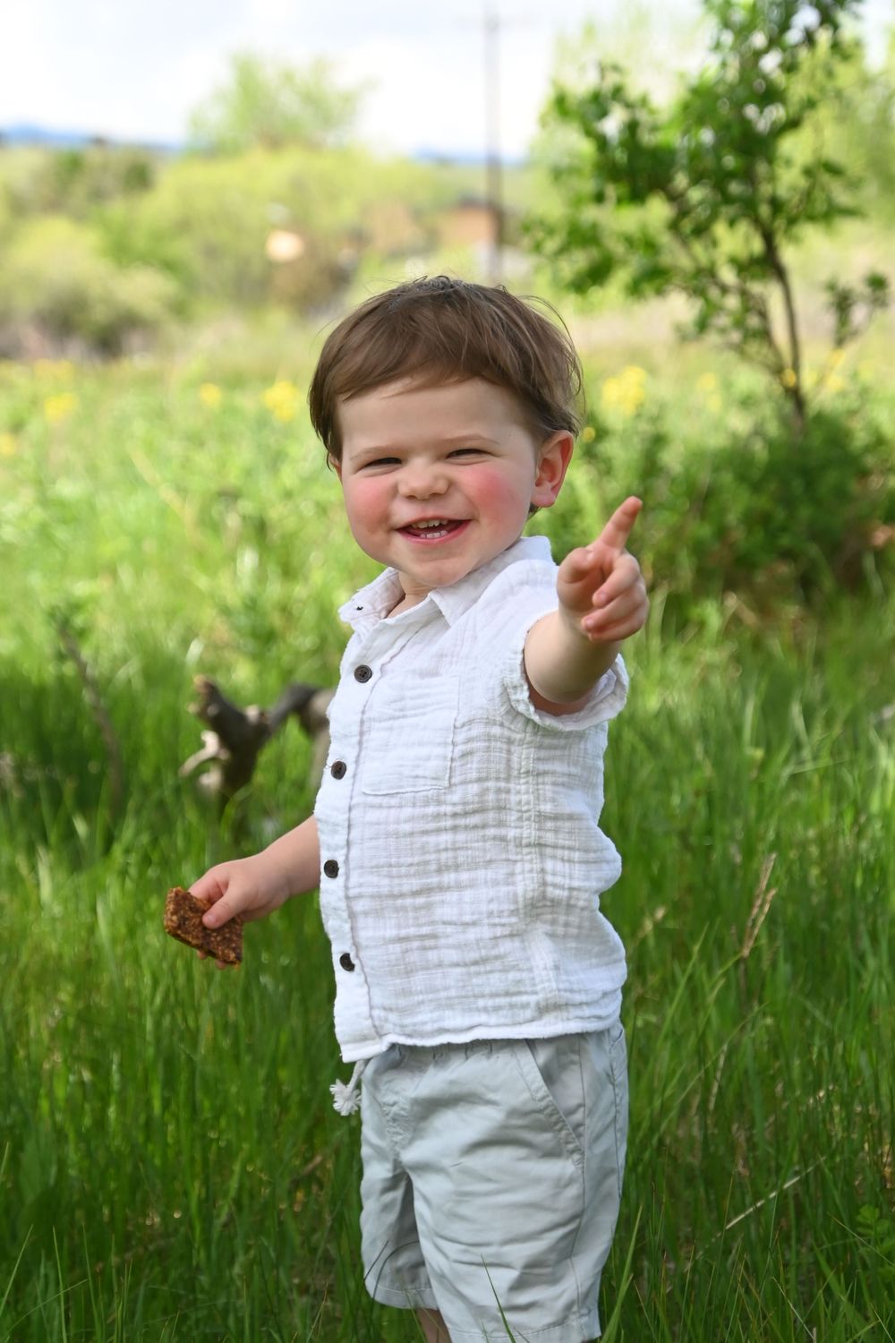 Small child in white button-down shirt and light pants pointing while standing in green grass on sunny day.