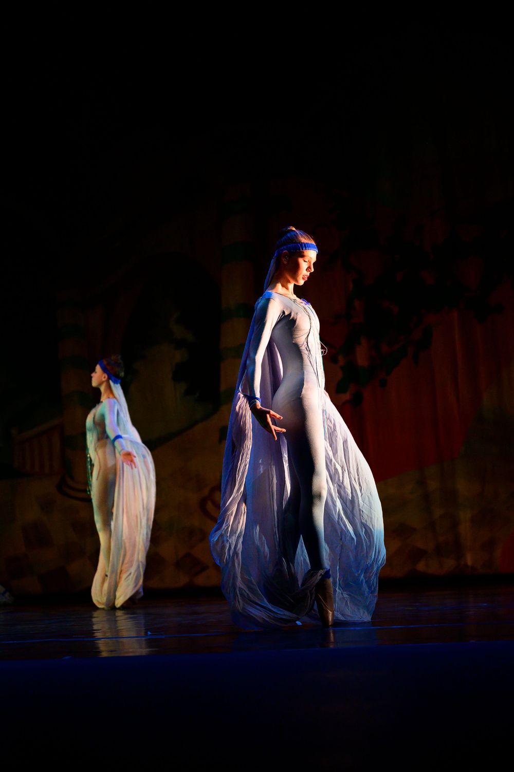 Two dancers in white flowing costumes performing on a dark stage with dramatic lighting effects.