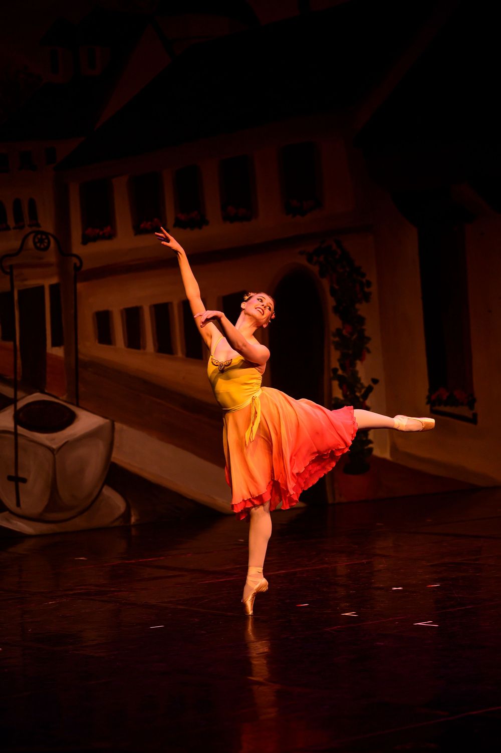 A dancer in an orange and red dress performs a dynamic contemporary dance sequence on a dimly lit stage.