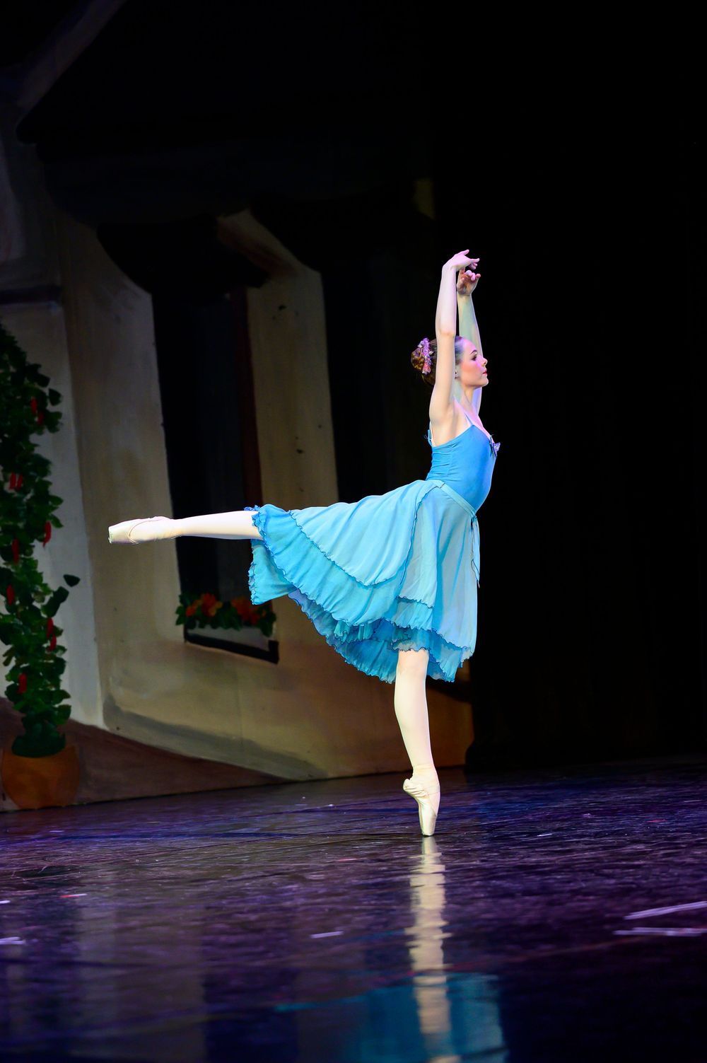 A ballet dancer in a flowing blue dress performs an arabesque pose on stage during a performance.