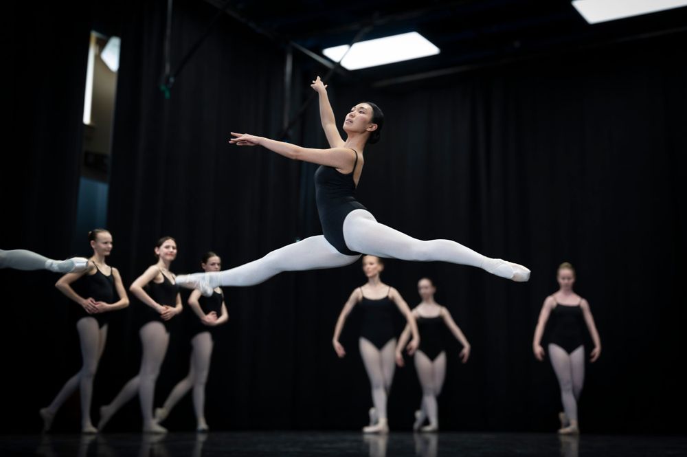 Ballet dancer leaping through air during practice with others in background.